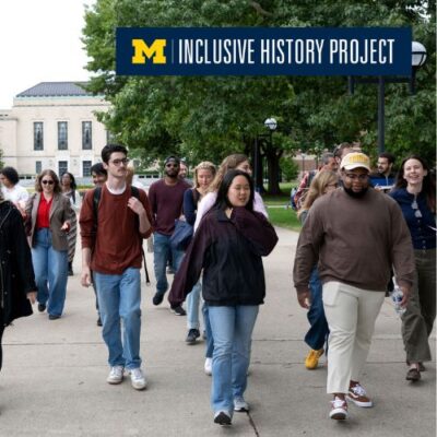 A group of people of varying nationalities walking on U-M's Central Campus in front of the Rackham building with the logo of the Inclusive History Project on the corner.