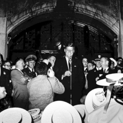 John F. Kennedy stands at a microphone surrounded by a crowd of people.