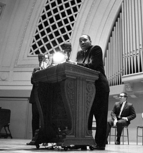 Martin Luther King Jr wears a dark suit and leans on a podium on a wide stage at the University of Michigan.
