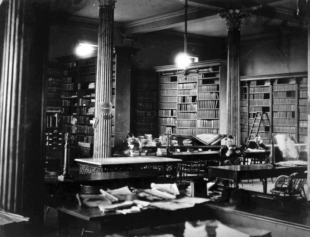 Black and white photo of a large room with tables and shelves of books, taken in the late 1800s. 