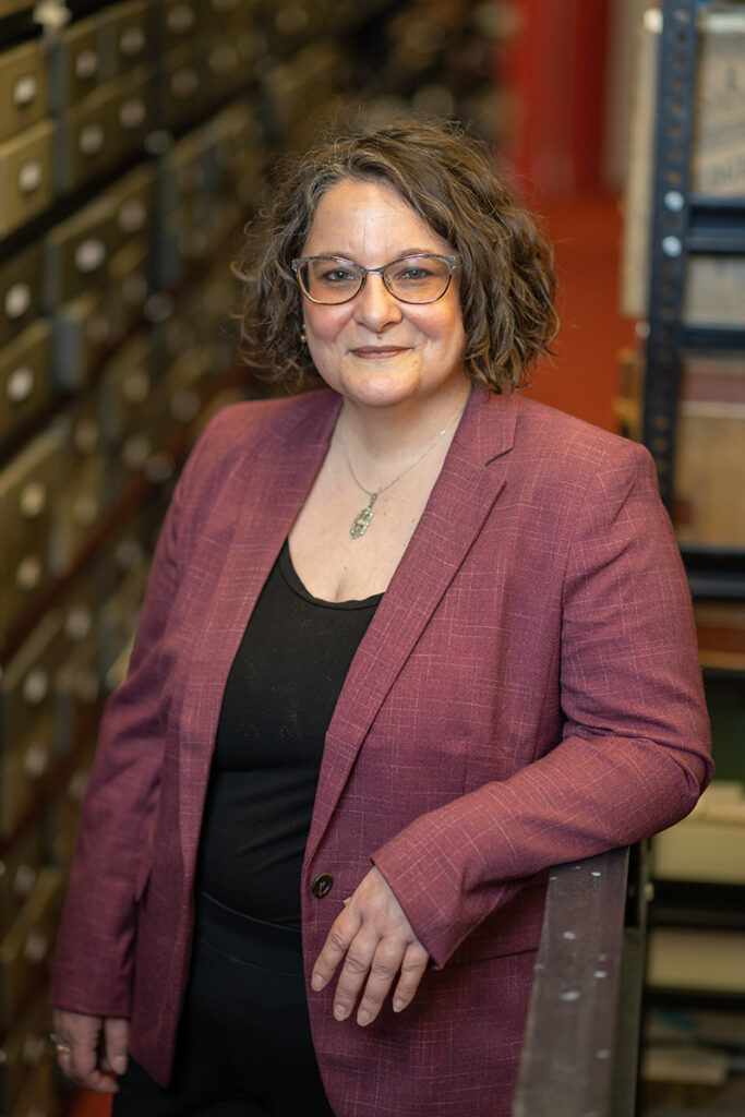 Woman wearing blazer and glasses with archived materials in background. 