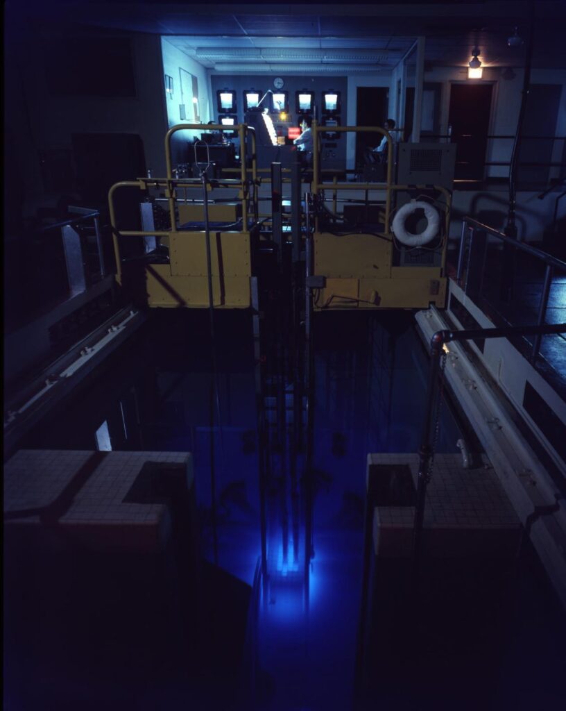 A glowing blue pool with a yellow "bridge" and lab above it. Glowing screens and technicians are visible in the lab. 