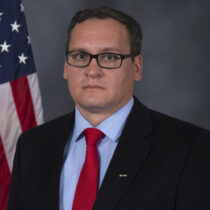 Man wearing glasses, a blue blazer and a red tie, with American flag in the background.