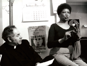 Man with priest collar sits on left looking at a woman on the right who is gesturing. 