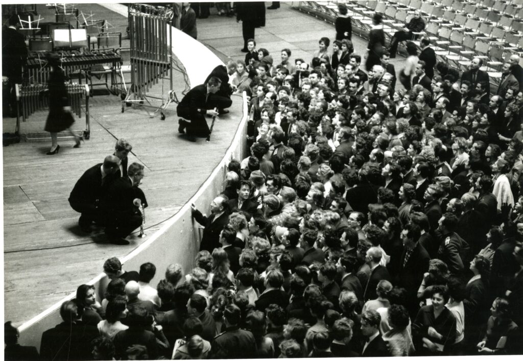 Black and white photo showing members of a large crowd pressing up against a stage where symphony band members kneel. 