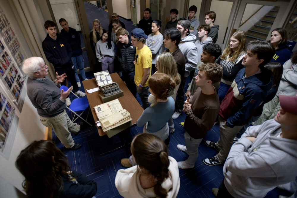 Man behind a table with scrapbooks surrounded by students from a U-M class.