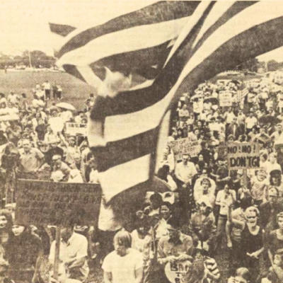 An American flag waves in front of a crowd of white protesters with signs.
