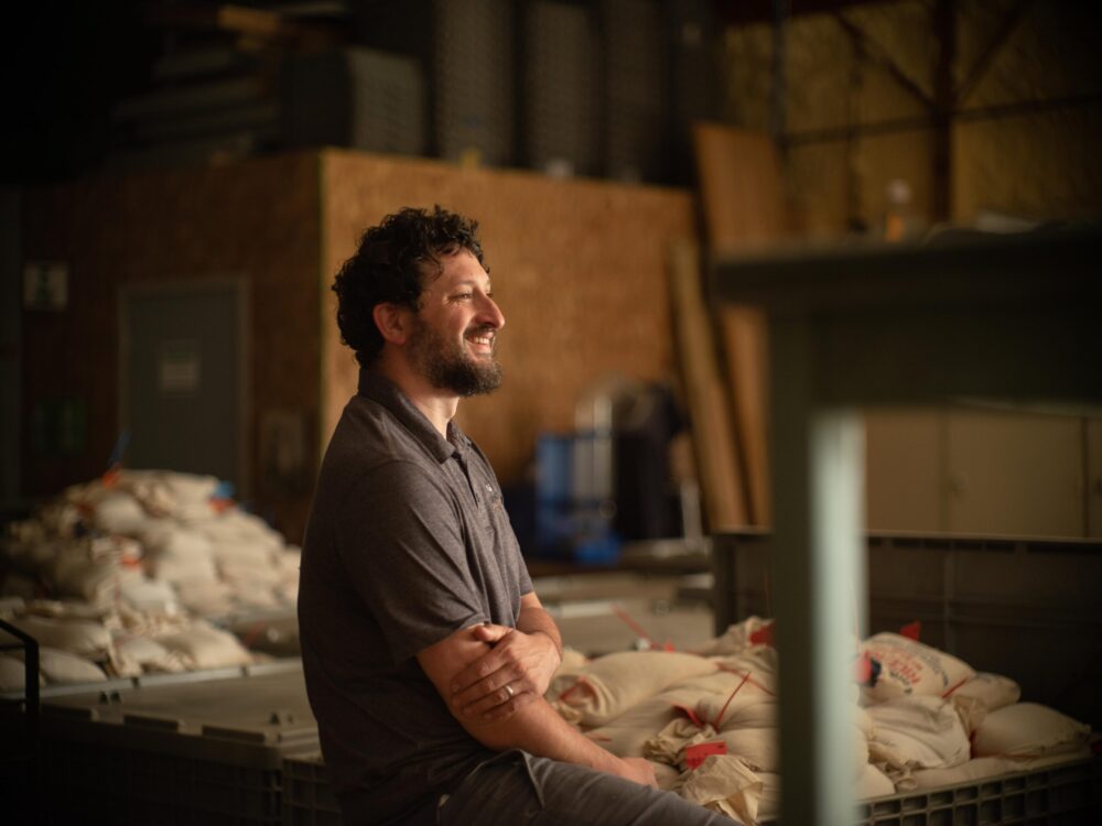 Ari Sussman wearing a grey t-shirt and standing in a warehouse with bags of rye seeds. 