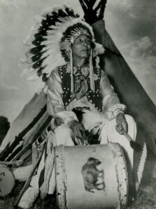 Native American man wearing a feather headdress and playing a drum. 