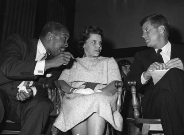 Woman in a dress is seated between two men who are talking. 
