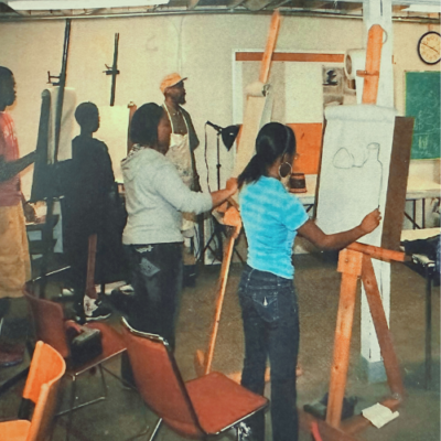 Individuals in a classroom stand at easels drawing on white paper.