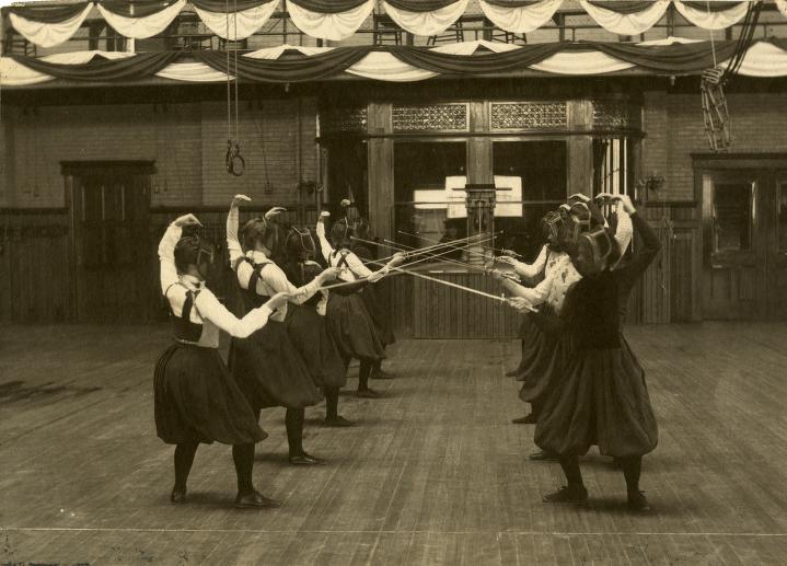 Women in dresses standing on a wooden floor with fencing swords in hand. 