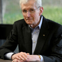 Man with white hair wearing a dark suit coat sits at a table.