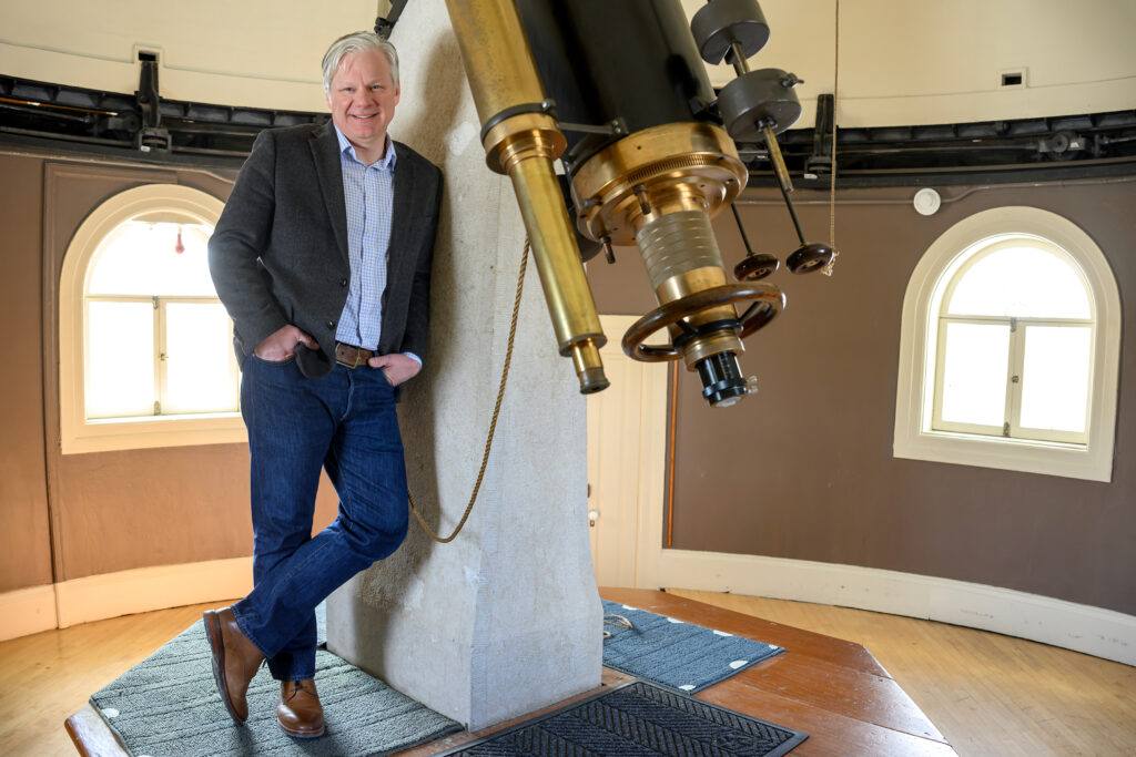 A man with hands in pockets wearing a collared shirt and sport coat leans against the foundation for a historic telescope. Rounded windows are in the background.