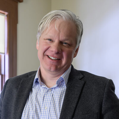 Man wearing collared shirt and a sport coat faces camera and smiles.