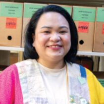 Researcher Eimee Lagrama wearing a brightly colored pink and yellow overshirt, and standing in front of boxes of historical materials.