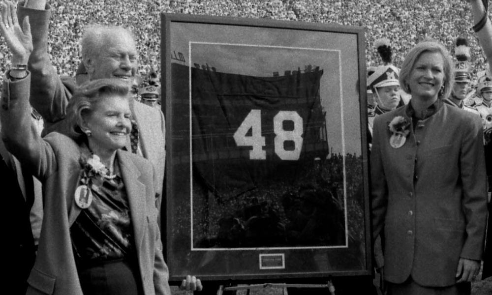 Gerald Ford's initial football number retirement ceremony in 1994, from the Kalmbach photographs, featuring Gerald Ford standing next to two women and a large framed "48" football jersey in front of a marching band on a football field.