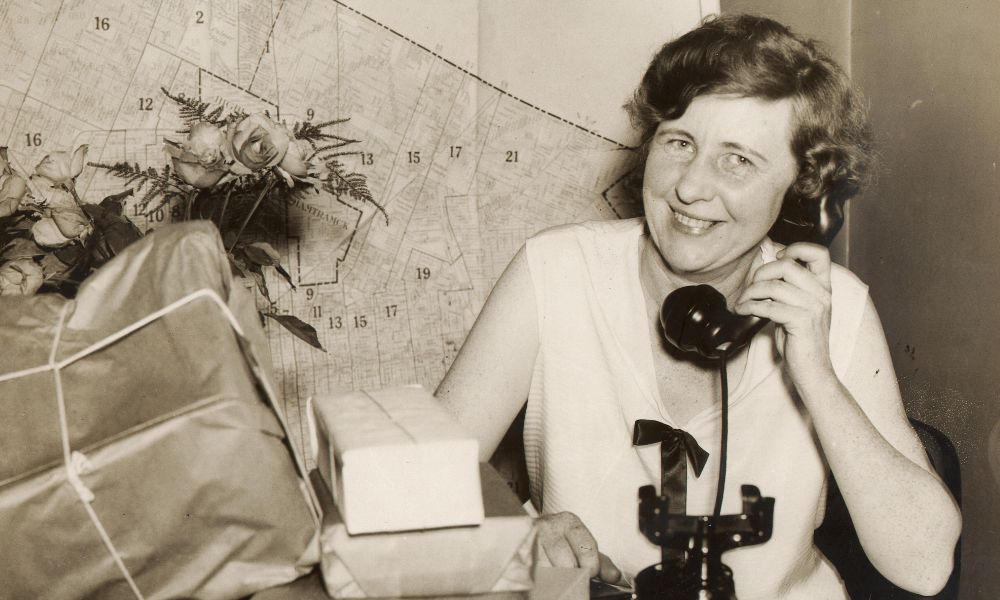 Josephine Gomon holding a telephone in front of a map of Detroit next to a pile of packages on a table.