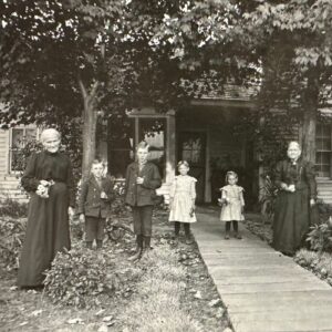 Two grandmothers and four children holding flowers they have just picked, standing in front of a house, with a walkway and trees leading up to it.