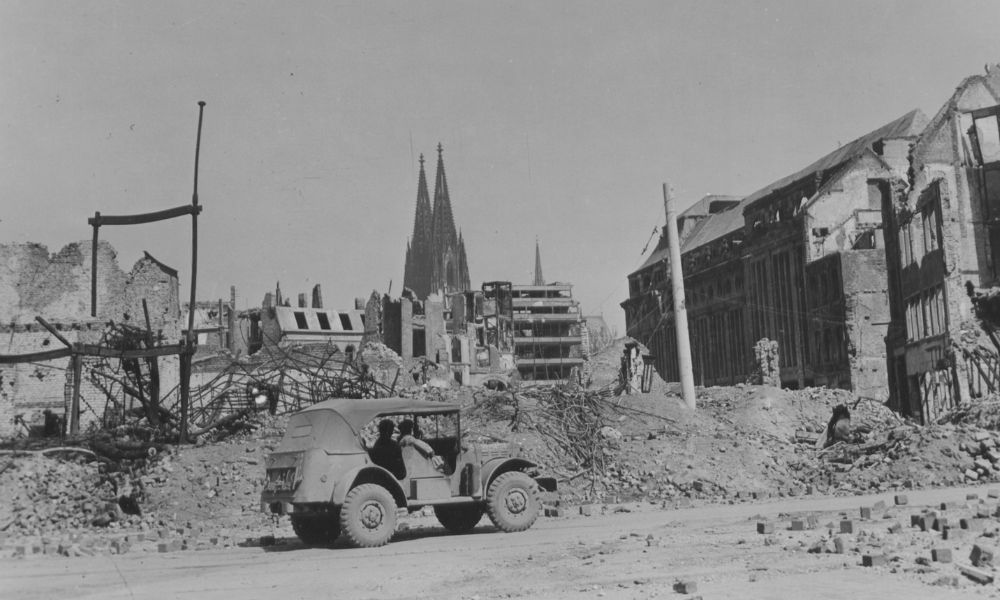 A military car drives through a street lined with the rubble of buildings in Cologne, Germany, during World War II.