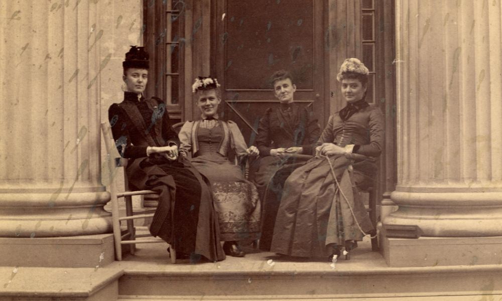 Alpha Phi sorority members at the University of Michigan, featuring four women sitting on the porch of a building with classical columns.
