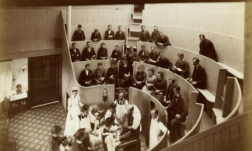 Homeopathic Amphitheater at the University of Michigan in 1893, featuring rows of students sitting inside an amphitheater, while doctors demonstrate a procedure on a patient.