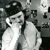 Jeep Holland holding a phone, wearing a headband and sunglasses on his head, with photos of his band behind him.