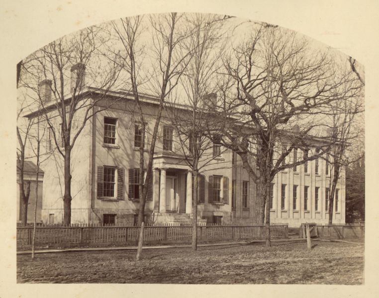 Southeast Professors' House, featuring a building with a small porch with two columns, an attached building with many windows, a grassy yard with trees, and a wooden fence.