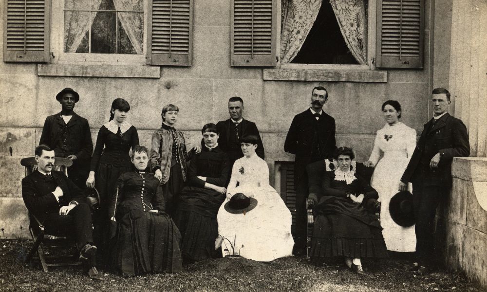 Staff of the Homeopathic Hospital in September of 1884, including men and women gathered outside of a building with lace curtains in the windows. Three women and one man sit in the front row, while three men and four women stand behind them. All are wearing fairly formal 1880s outfits, with the men wearing suits, and the women wearing gowns. 