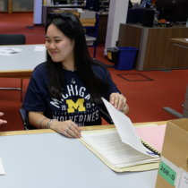 Woman with dark hair and blue shirt sits at a table with a folder and box in front of her.