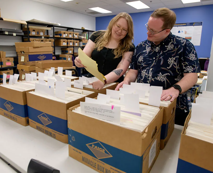 A woman holds a yellow sheet of paper at arm's length. To her right, a man inclines his head to look at it. In front of them are many brown archival boxes filled with white folders.
