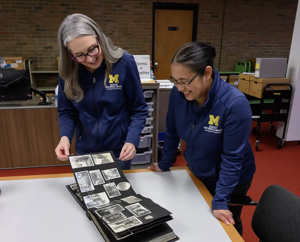 Two women wearing blue jackets with a yellow Block M study the pages of a scrapbook on a table in the Bentley's reading room. The scrapbook pages are dark and have old black-and-white photos glued to them.