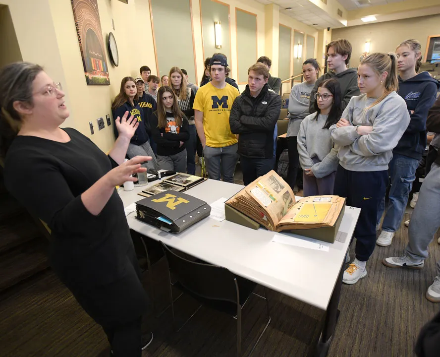 A woman in a black dress gestures with her hands. A large group of students from a sports history class gathers around, listening to her talk. On a white table in front of her is a blue scrapbook with a yellow Block M on the cover.