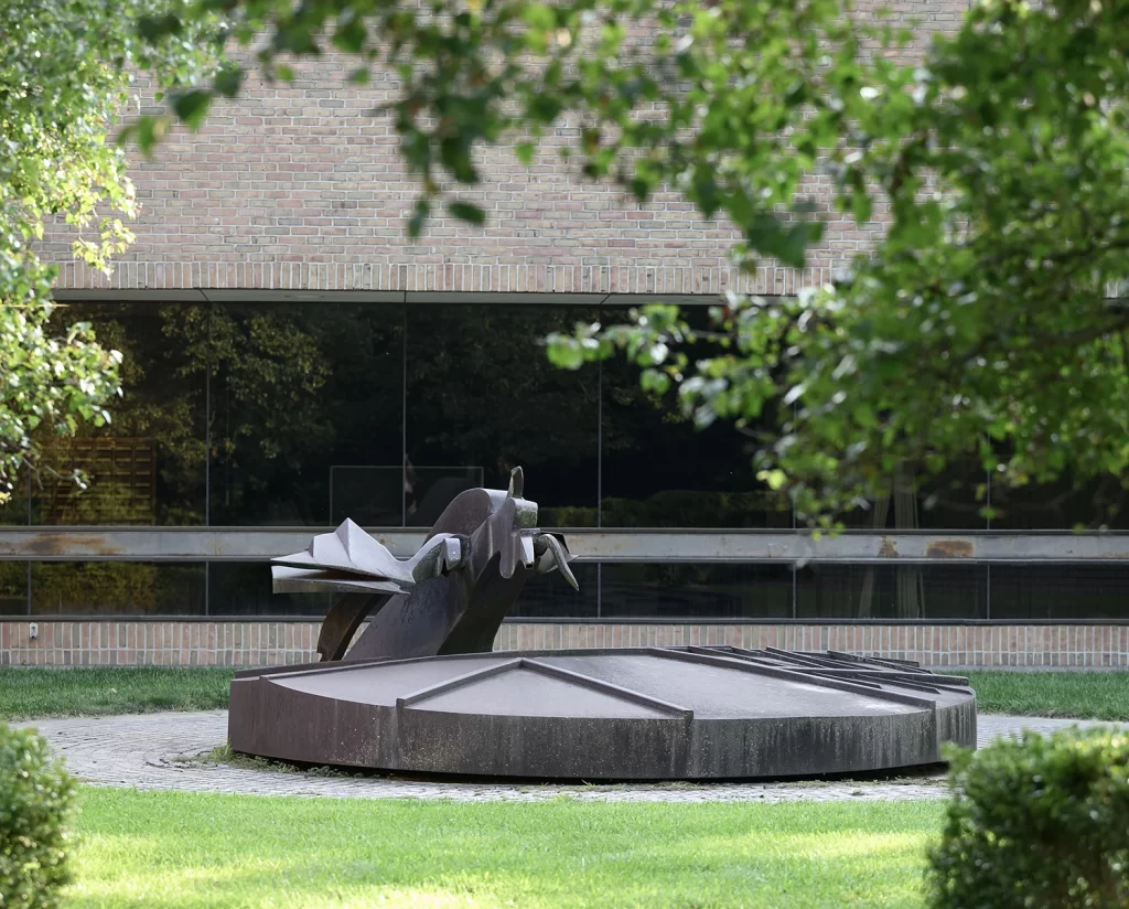 A large metal sculpture with a round base is framed by green tree branches and grass. Behind it are windows to the Bentley Historical Library's reading room.