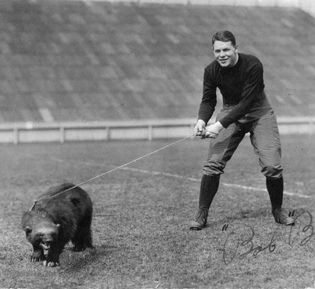A black and white photos shows a man wearing long pants, boots, and a dark shirt posing with a stuffed wolverine on the University of Michigan football field.
