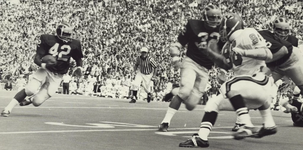 A historical football photo featuring the University of Michigan football team competing in a game, running down the field, while a crowd looks on from the stands inside a stadium.