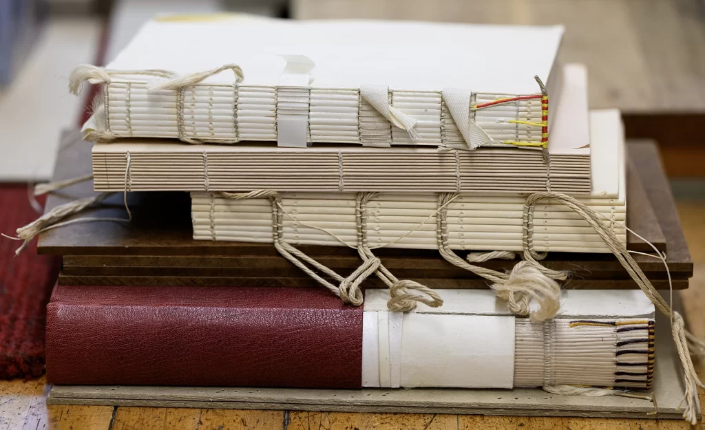 A stack of partially bound books in the Conservation Lab at the Bentley Historical Library, featuring book blocks with sewn spines exposed to show threads, ribbon, and thin rope, awaiting book covers.