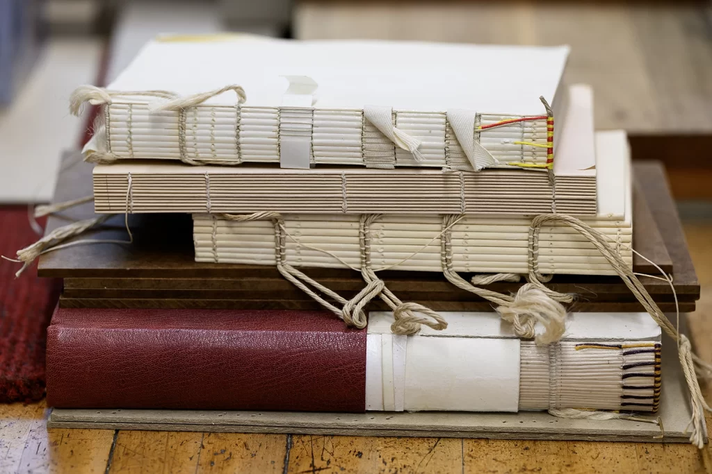 A stack of partially bound books in the Conservation Lab at the Bentley Historical Library, featuring book blocks with sewn spines exposed to show threads, ribbon, and thin rope, awaiting book covers.