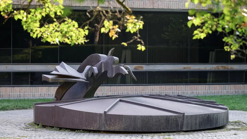 The "Historical Circle" statue by Richard Hunt in the Bentley Historical Library's garden courtyard, featuring a welded steel abstract statue of a wavy line connected to a circle that looks like a sundial, with a brick building and large glass windows behind it, a grassy lawn, and leafy tree branches in the upper edge of the photo.