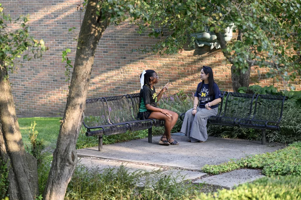 Two women seated on benches in a courtyard, facing each other and talking. A brick wall is behind them, with trees and grass on either side.