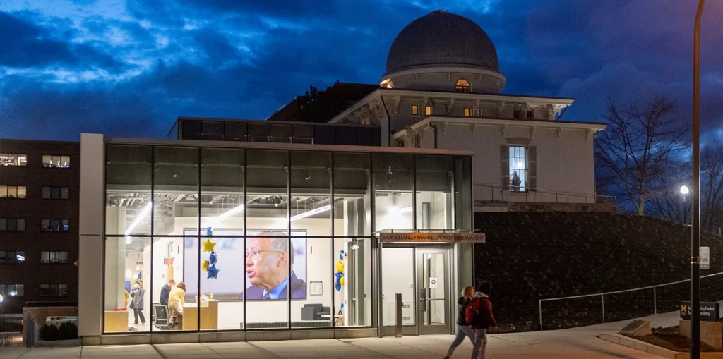 The Judy & Stanley Frankel Detroit Observatory building, featuring a historical building with an observatory dome, and the newer addition with glass paneling, illuminated at night, on U-M's Central Campus in Ann Arbor.
