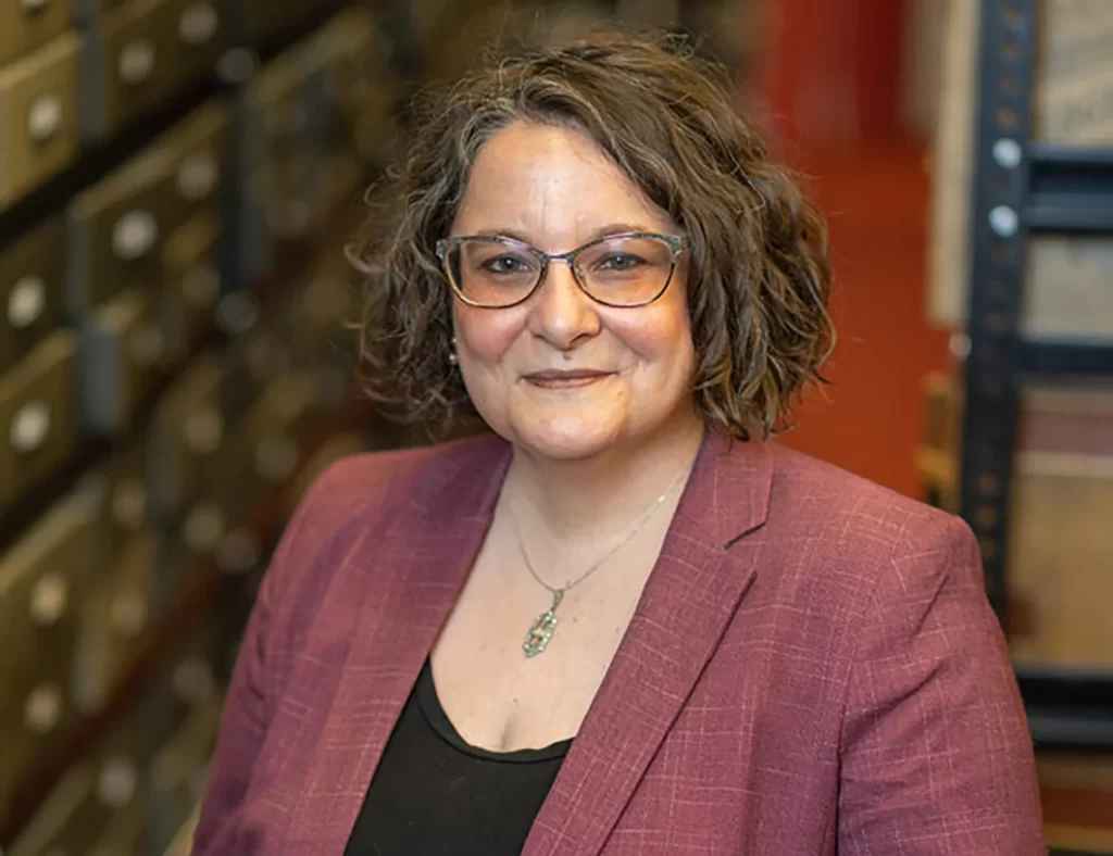 Bentley Director Alexis Antracoli stands in the Bentley Library stacks, with boxes and books in the background. She is wearing a mauve suit jacket and has dark curly hair and glasses.