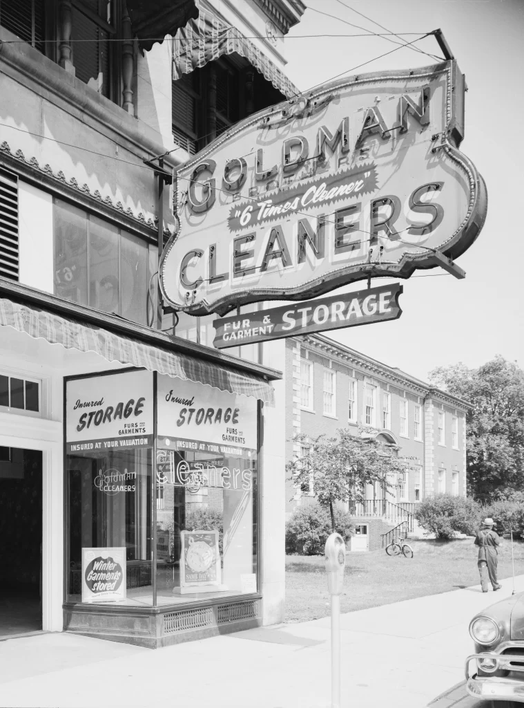 A black and white photo of a storefront exterior. A large sign says "Goldman Cleaners." In front of the store is a sidewalk. Partially visible is a parking meter and old car.