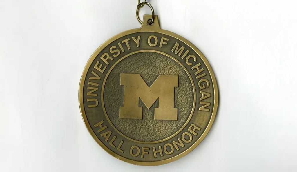 A bronze-colored round medal with a Block M in the middle says "University of Michigan" at the top and "Hall of Honor" on the bottom.