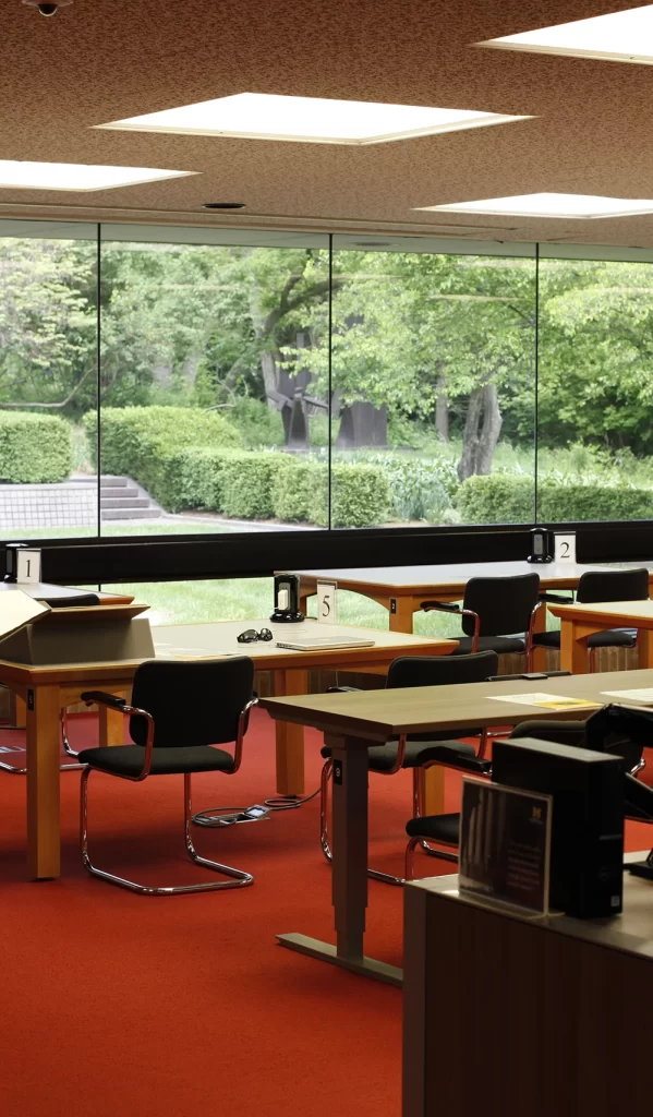 Several desks with black chairs in the Bentley Historical Library's reading room sit in front of large windows facing a green courtyard.