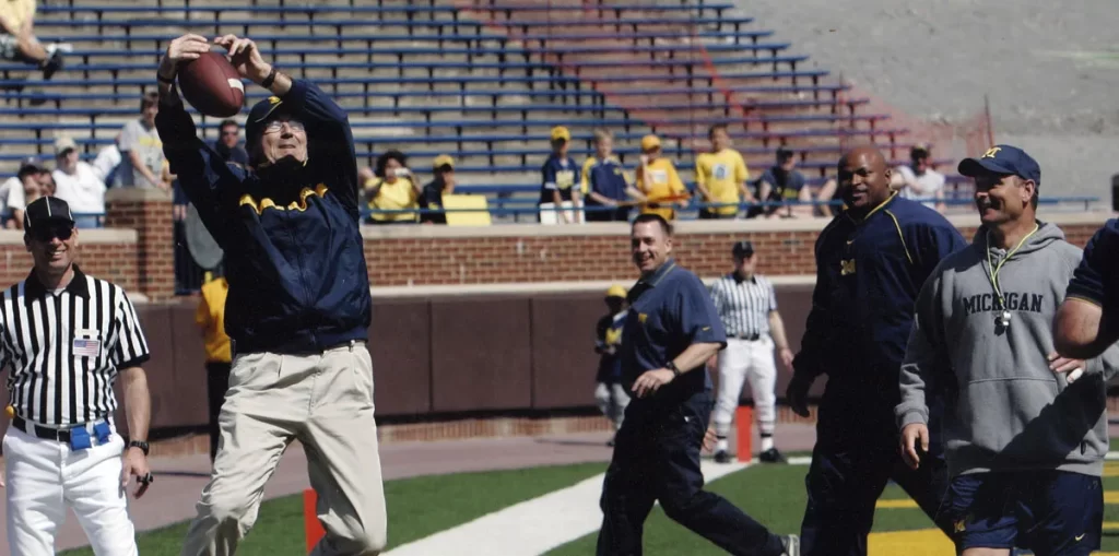Football coach Lloyd Carr, wearing khakis and a blue jacket, reaches up to catch a football pass. He is on green turf at the University of Michigan football stadium, surrounded by several other people including a referee in a black and white striped shirt and men wearing Michigan shirts.