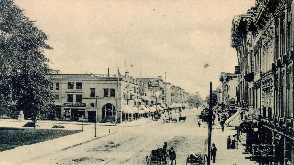 Main Street in Ann Arbor in 1904, featuring tall buildings with awnings and decorative brickwork, trees, telephone poles, horse-drawn carts on the road, and pedestrians walking on the sidewalks.