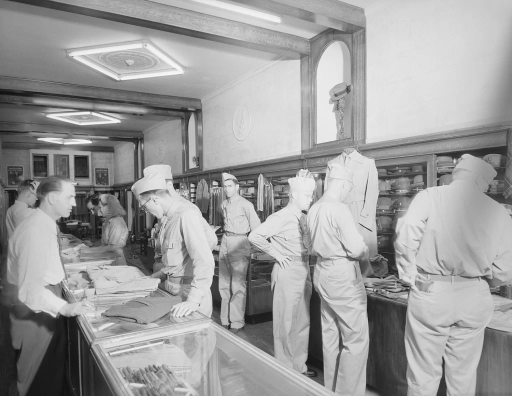 A black and white photo of men in military uniforms shopping in a store. A man behind the counter wearing a white shirt and dark pants waits on them.