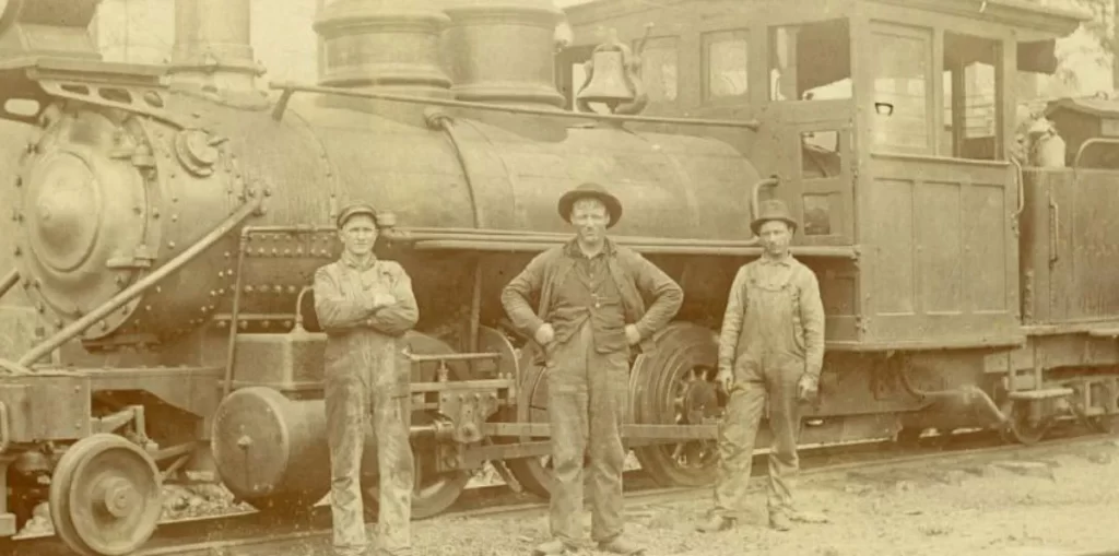 Three men stand in front of a large train engine in a black and white photo from the late 1800s.