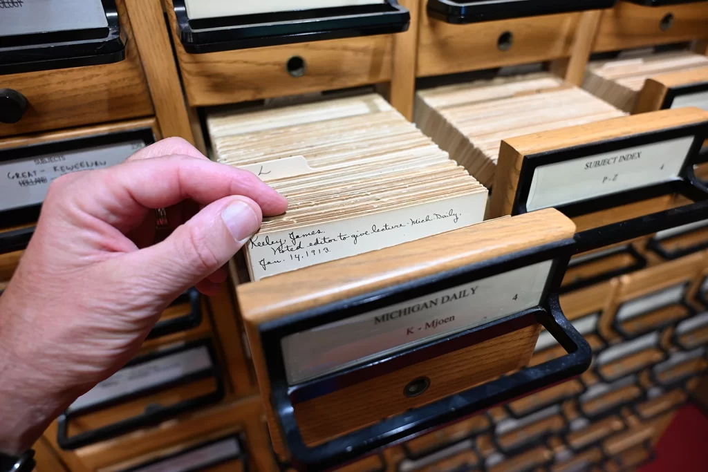 A small card catalog drawer is pulled open, and a library patron browses the cards. Only the patron's hand is visible.